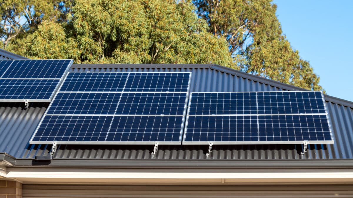 Rooftop solar panels installed on a corrugated metal roof, with trees and blue sky in the background.